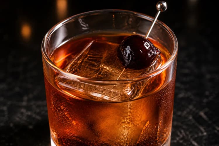 Little Italy served in a rocks glass over a large ice cube, dark amber in color, garnished with a cherry.