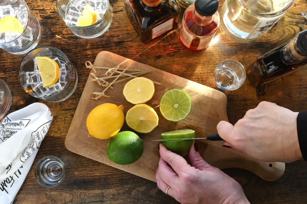 Overhead flat lay of citrus, spirits, and ice while slicing lime for cocktails