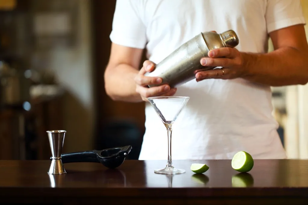 Home bartender with a cocktail shaker, jigger, and lime on a wooden counter