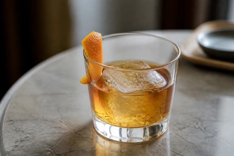 Añejo Old Fashioned in a rocks glass over a large ice cube, deep gold, orange peel.