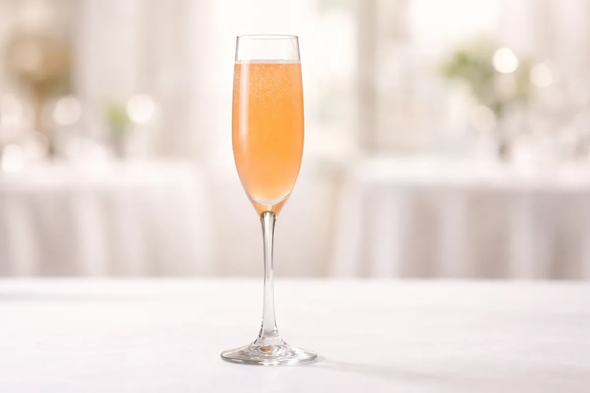 A Bellini cocktail in a tall Champagne flute with pale peach color and fine bubbles on a white tablecloth.