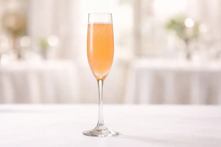 A Bellini cocktail in a tall Champagne flute with pale peach color and fine bubbles on a white tablecloth.