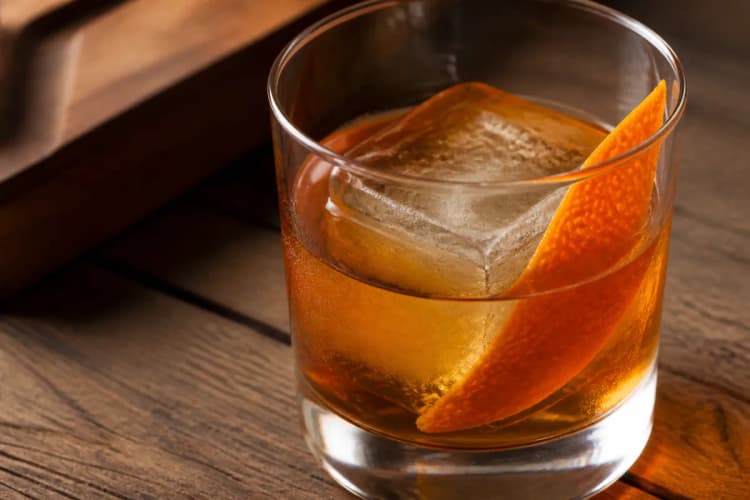 Maple Old Fashioned in a rocks glass over a large ice cube, deep amber, orange peel.
